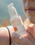 woman holding skincare bottle in front of her shoulder to the side of her face.