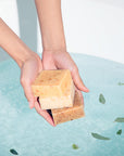 Hand cupping three LATHER soap bars over a bathtub filled with clear blue water