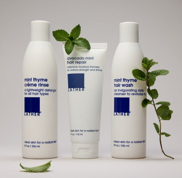 Three white Lather haircare products on a light gray background: a mint thyme crème rinse bottle, an avocado mint hair repair tube, and a mint thyme hair wash bottle, styled with mint leaves.