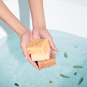 Model hands holding three stacked bars of LATHER soap over a bathtub full of clear blue water. 
